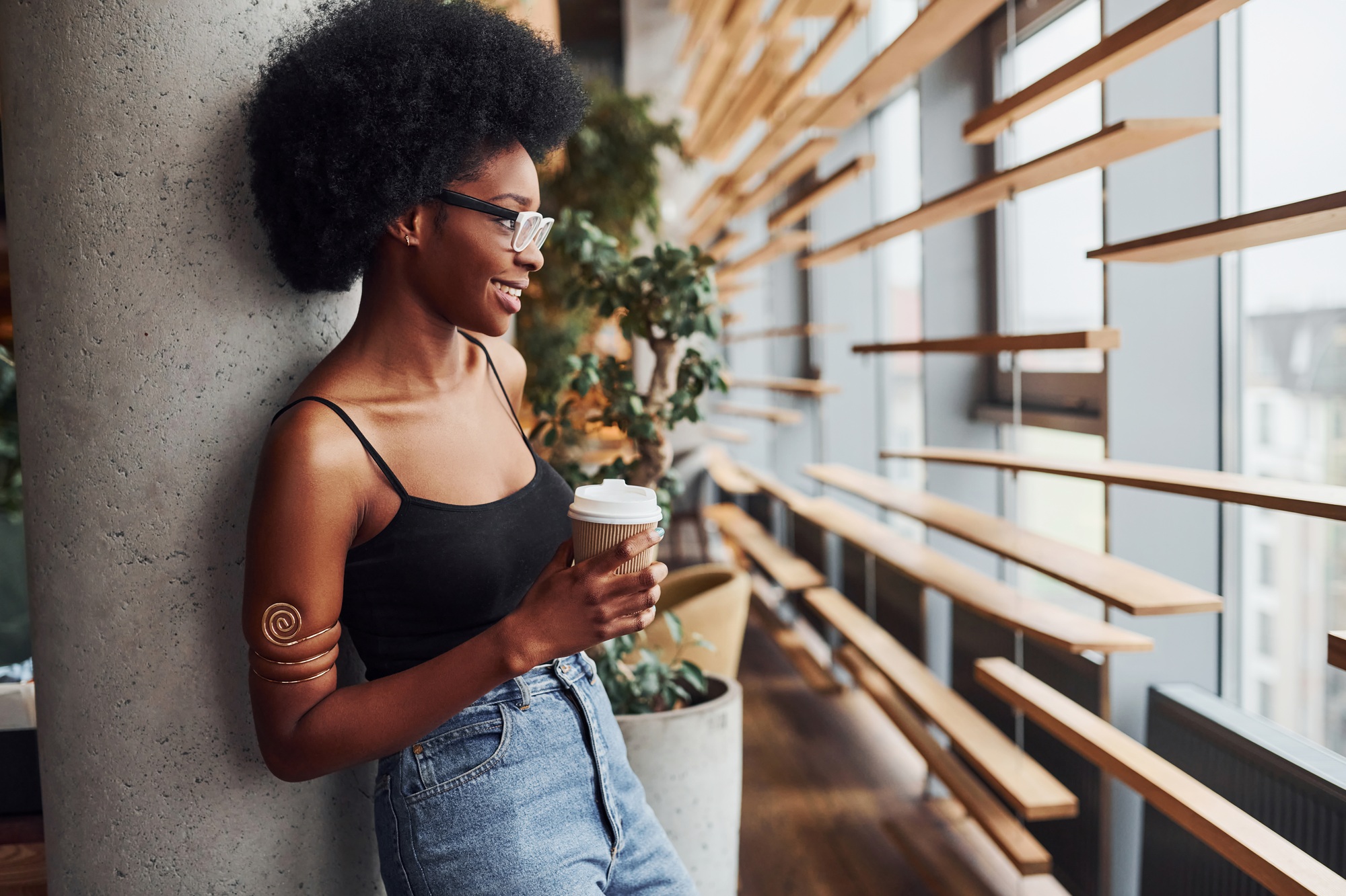 African girl with curly black hair and in casual clothes standing indoors with cup of drink